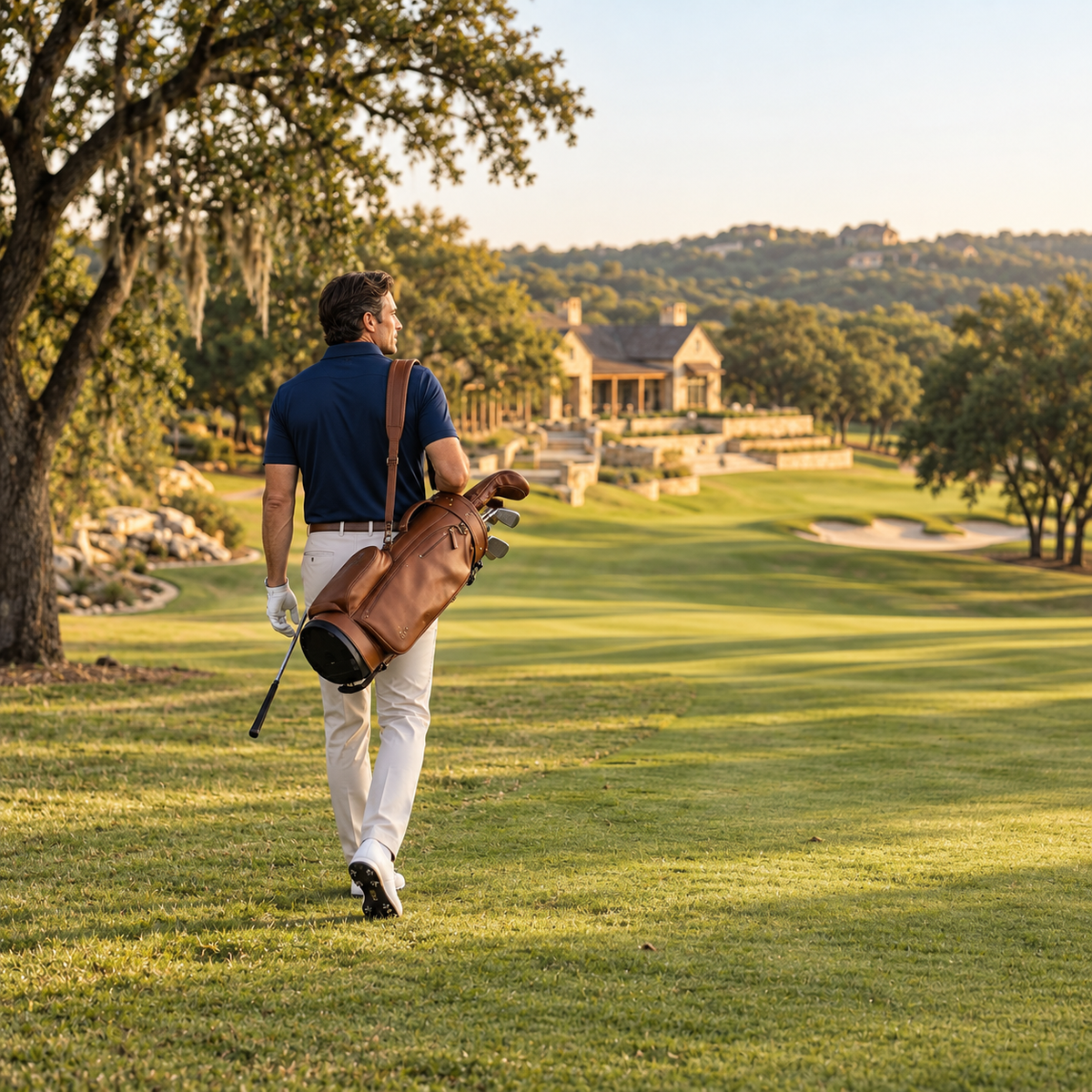 Golfer wearing a navy Ringo Reiner polo and light trousers walks along a sunlit fairway carrying a brown leather golf bag.